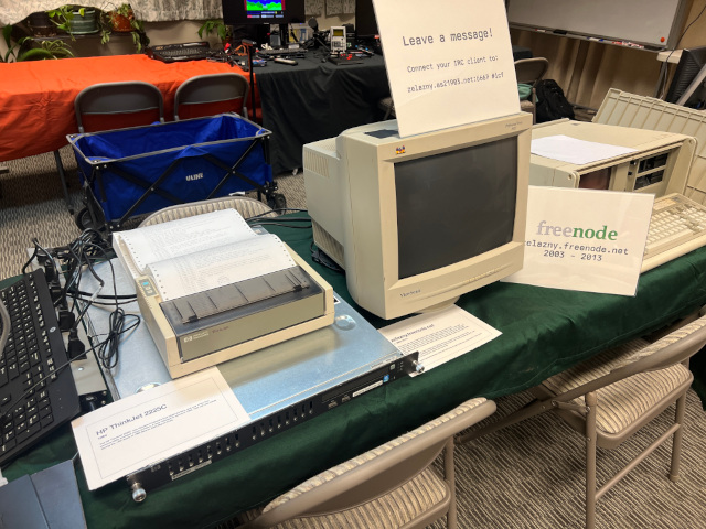 A number of items on a table in a room. From left to right, there’s a server with a printer on top of it, a CRT monitor and an IBM 5155 Portable Personal Computer
